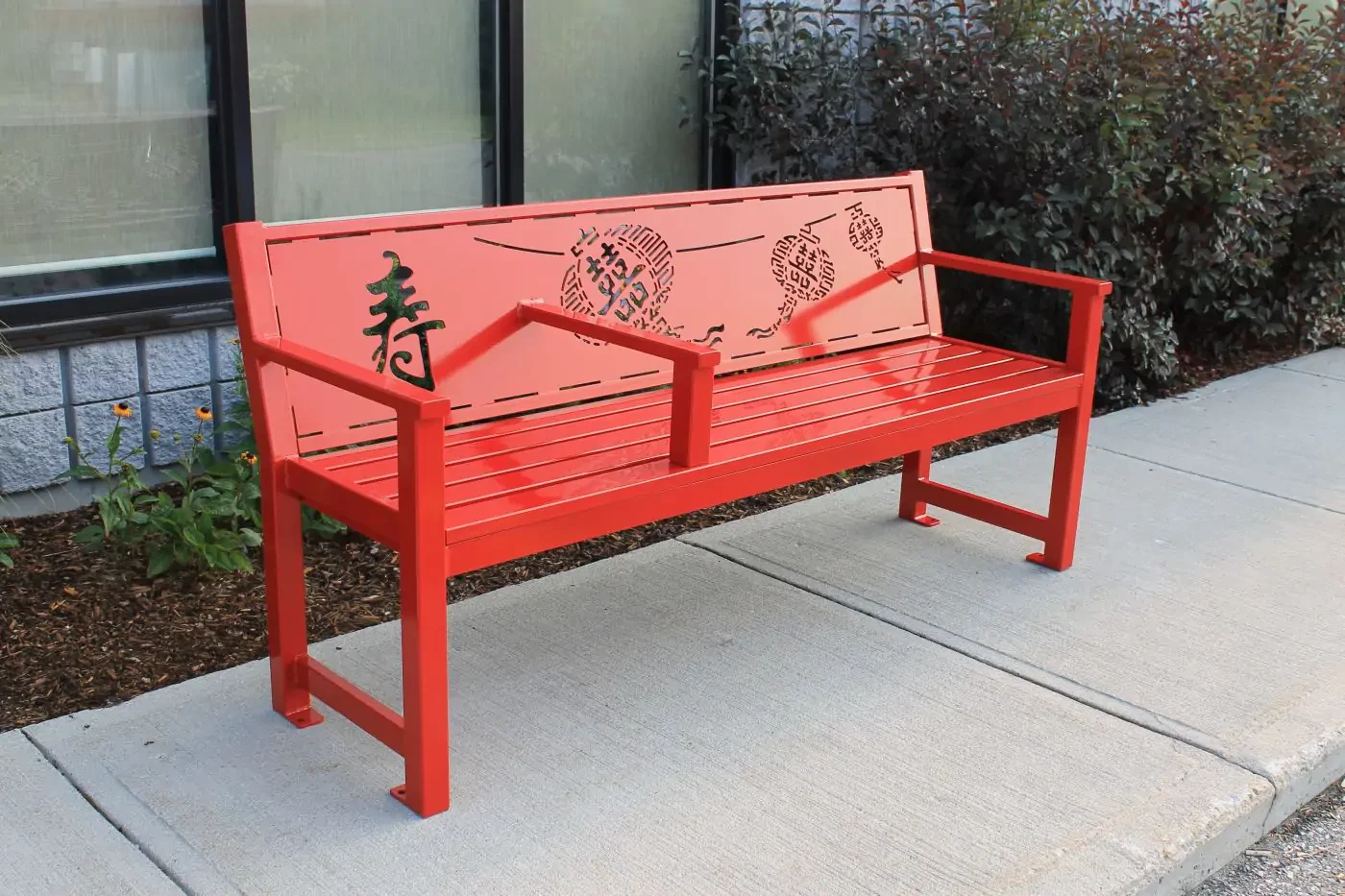 A bright orange metal bench with Asian-inspired cutout designs and a backrest, placed on a sidewalk near a building with bushes behind it.