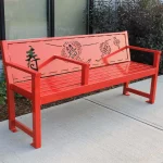 A bright orange metal bench with Asian-inspired cutout designs and a backrest, placed on a sidewalk near a building with bushes behind it.