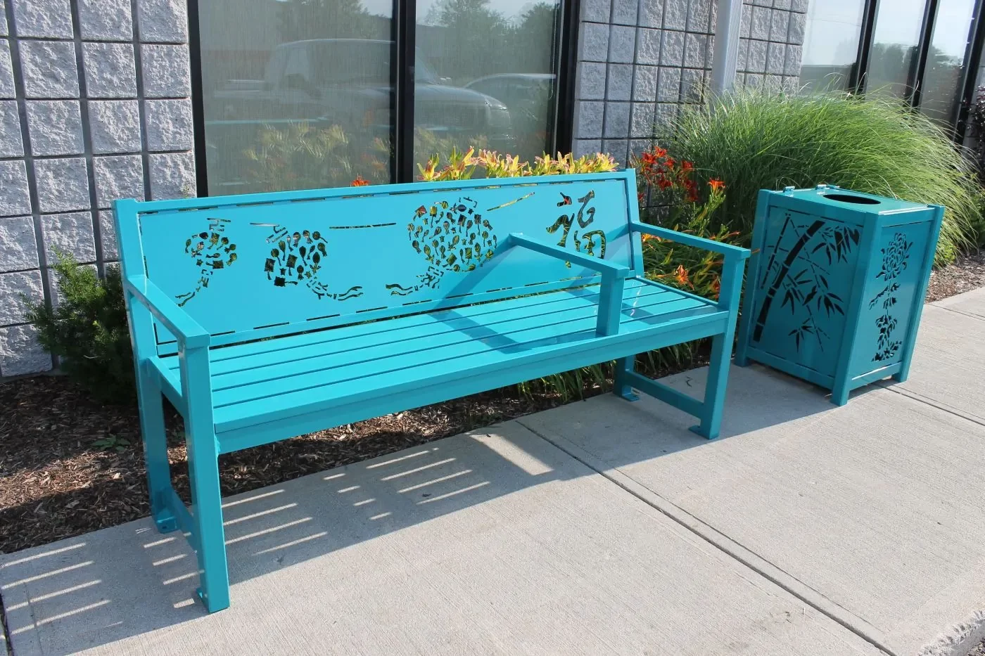 A turquoise metal bench with decorative cutouts and a matching trash can is placed on a sidewalk beside a building and landscaping.