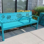 A turquoise metal bench with decorative cutouts and a matching trash can is placed on a sidewalk beside a building and landscaping.