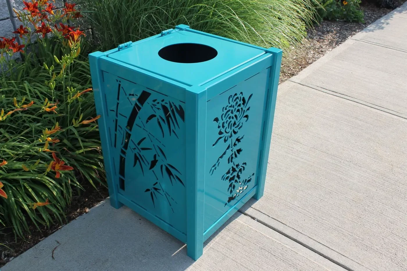 A teal metal trash bin with decorative cutouts stands on a concrete sidewalk next to green plants and orange flowers.