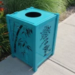 A teal metal trash bin with decorative cutouts stands on a concrete sidewalk next to green plants and orange flowers.