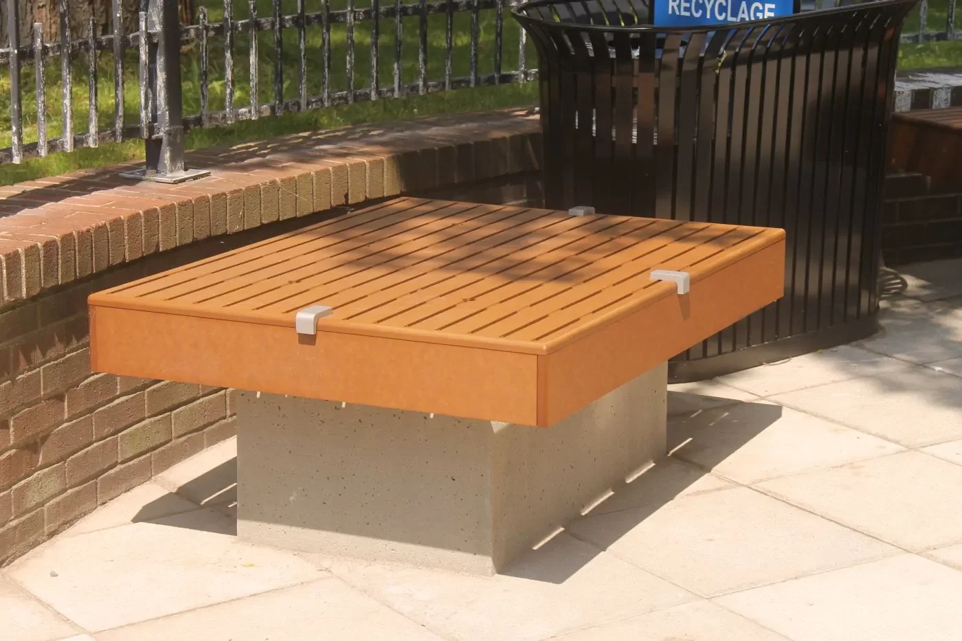 ON_VILLE-DE-GATINEAU-2 A modern wooden bench with a concrete base is placed on a paved area near a brick wall and a black recycling bin.