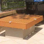 A modern wooden bench with a concrete base is placed on a paved area near a brick wall and a black recycling bin.