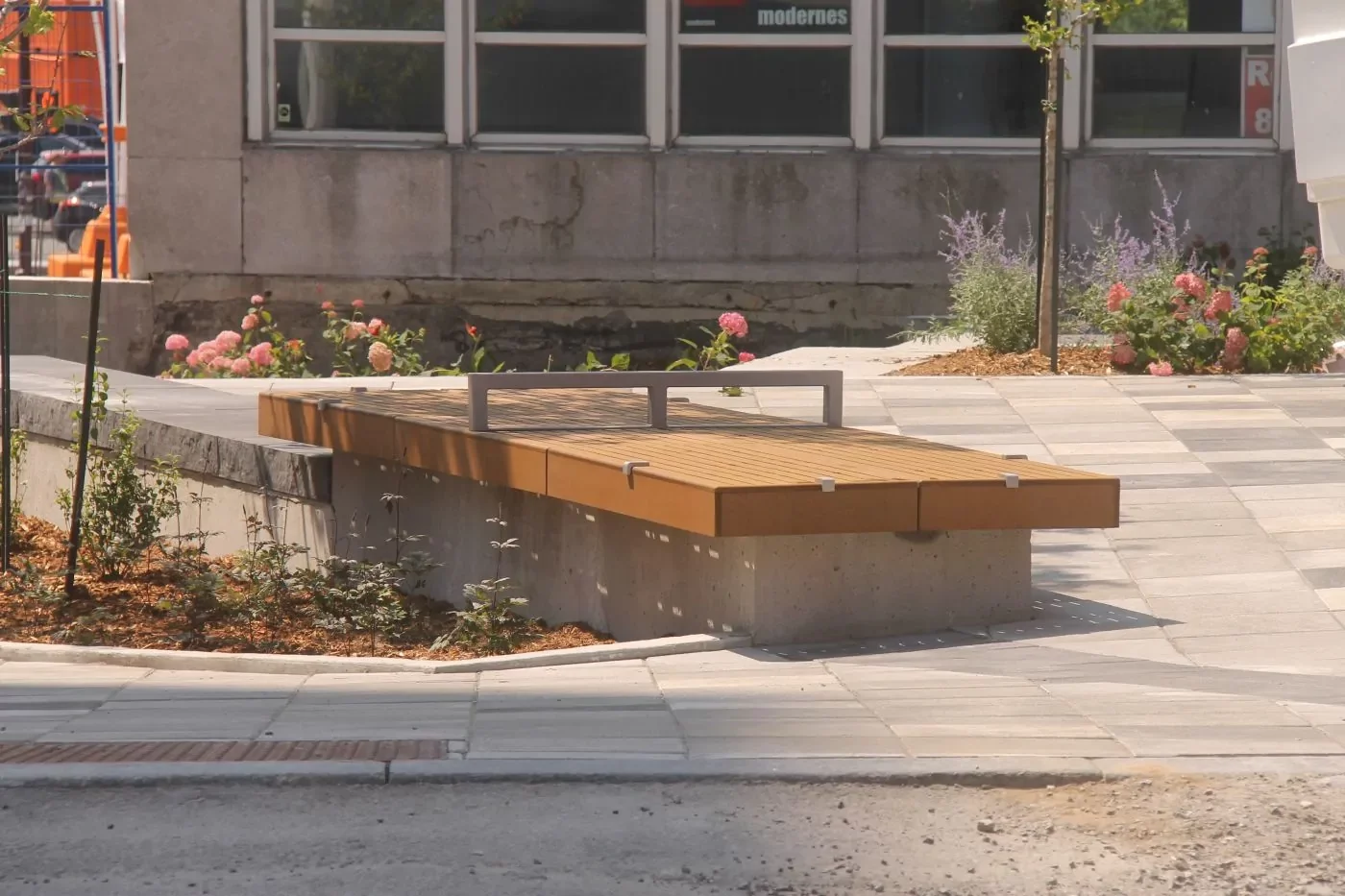 A wooden bench with a metal bar across the middle is situated on a paved area near a building and landscaped flower beds.