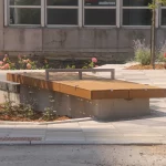 A wooden bench with a metal bar across the middle is situated on a paved area near a building and landscaped flower beds.