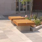 Two wooden benches with concrete bases are placed beside a small garden bed with flowers and young trees on a tiled outdoor area.