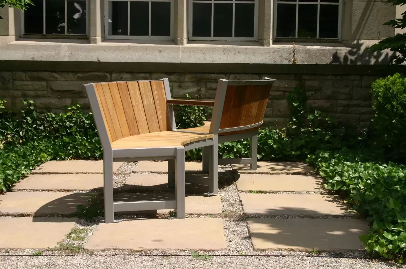 Two wooden chairs with metal frames are joined together at a right angle on a stone patio, set against a building with windows and surrounded by greenery.