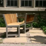 Two wooden chairs with metal frames are joined together at a right angle on a stone patio, set against a building with windows and surrounded by greenery.
