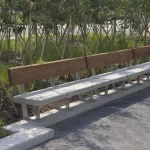 A long outdoor bench with a concrete base and wooden backrest is positioned beside a row of young trees in a landscaped area.