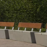 Three modern benches with wooden backs and concrete bases are arranged along a paved walkway with green foliage in the background.