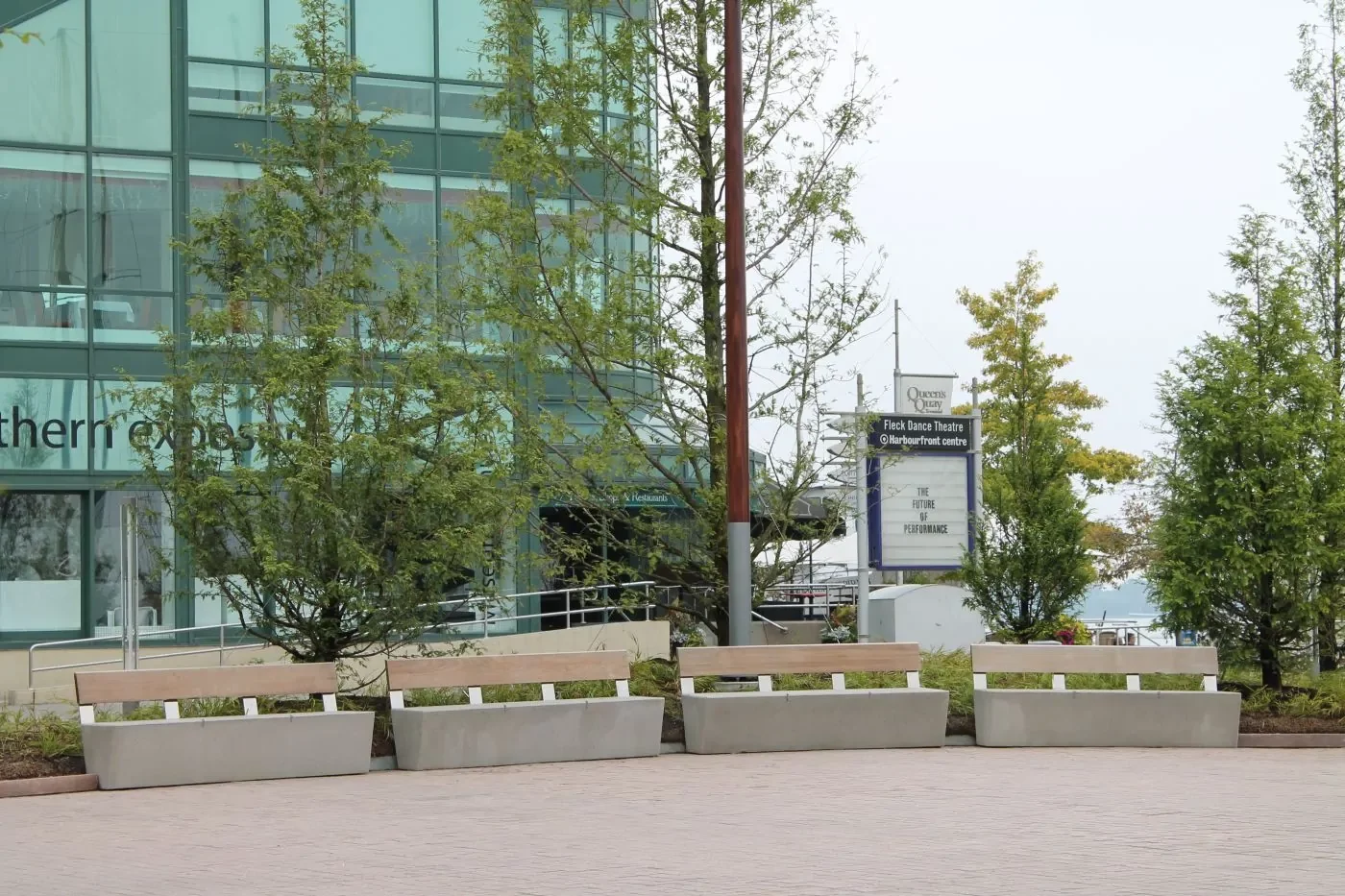 Concrete benches and large rectangular planters with trees are arranged outside a modern glass building; a sign for a theatre is visible in the background.