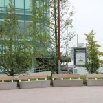 Concrete benches and large rectangular planters with trees are arranged outside a modern glass building; a sign for a theatre is visible in the background.