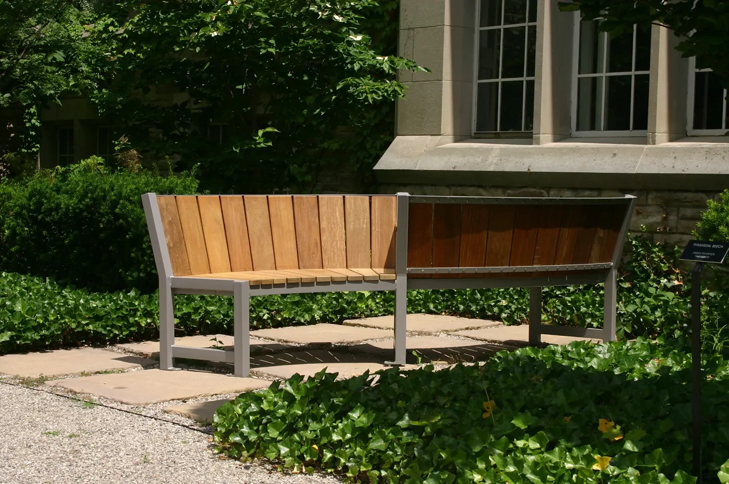 A wooden bench with one half made of light wood and the other half made of dark wood sits in a garden area outside a building with large windows.