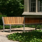 A wooden bench with one half made of light wood and the other half made of dark wood sits in a garden area outside a building with large windows.