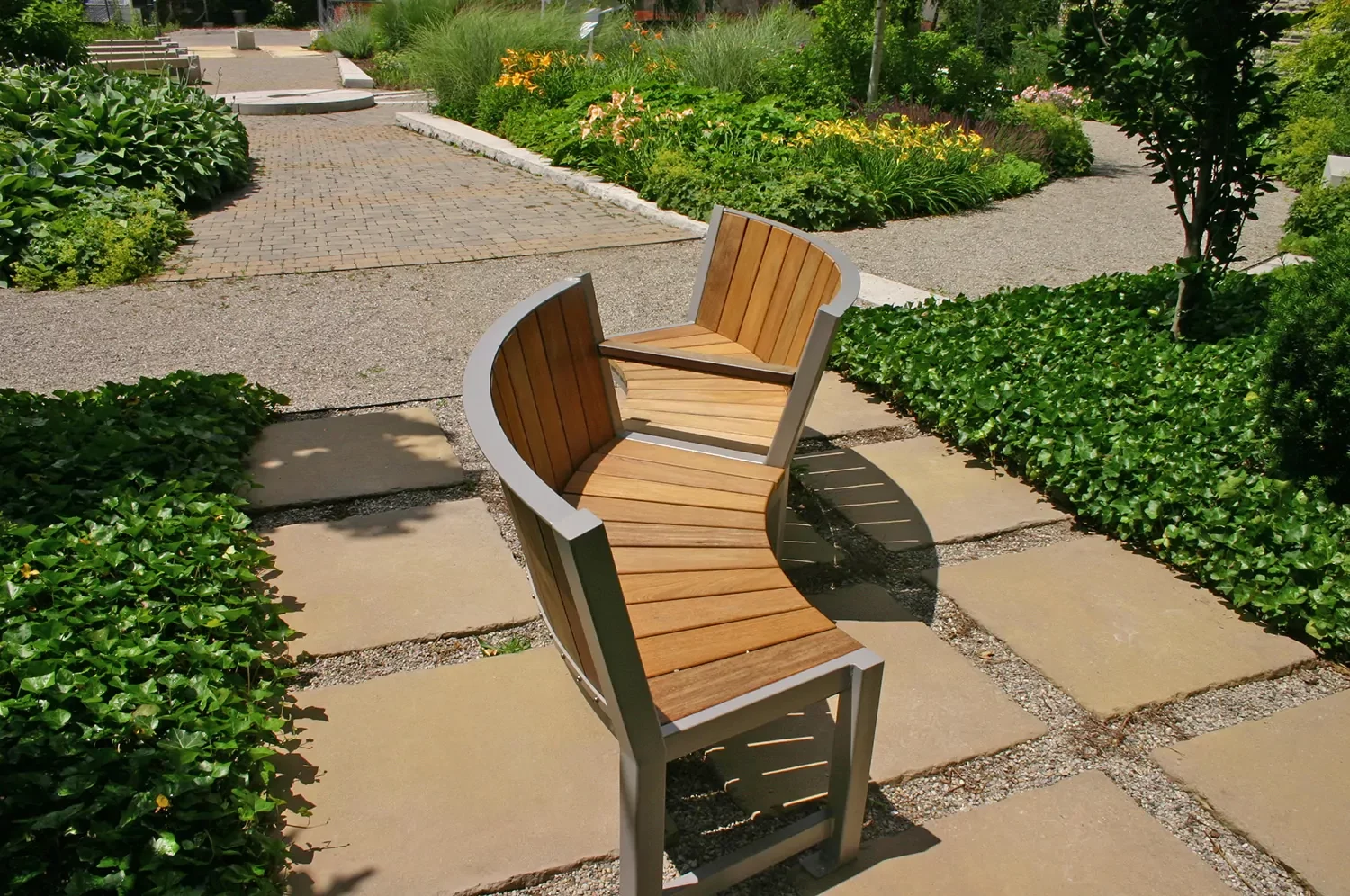 Two curved wooden benches with metal frames are positioned back-to-back on a stone path in a landscaped garden with greenery and flowering plants.