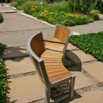 Two curved wooden benches with metal frames are positioned back-to-back on a stone path in a landscaped garden with greenery and flowering plants.