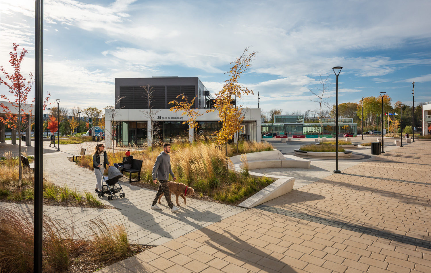A child sits on a Maglin Ogden Bench while a woman walks by with a stroller and a man walks a poodle-type dog on a pathway in Agora Plaza, Gatineau, QC. There is plentiful outdoor seating for comfortable use outdoors.