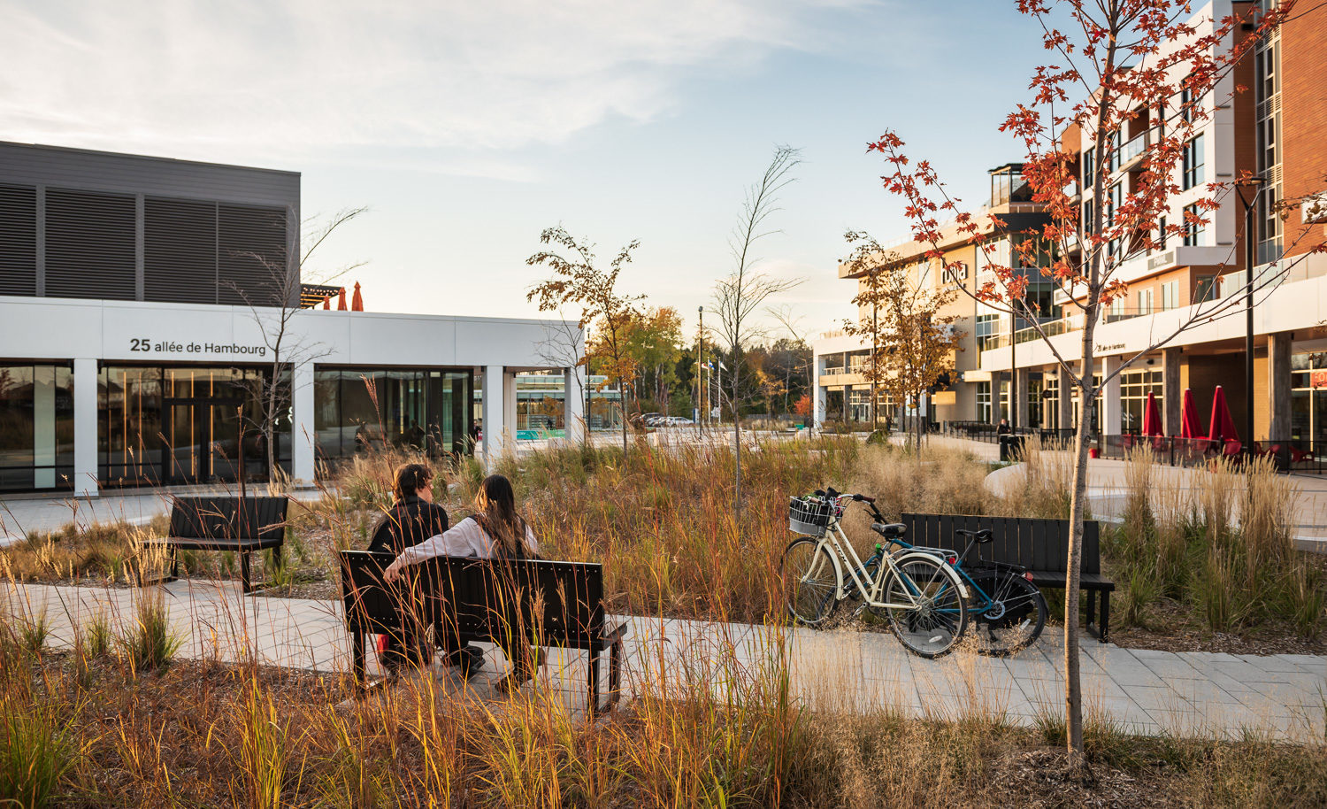 A couple sits on a Maglin Ogden Bench on a paved pathway in AGORA Plaza in Gatineau. There bikes are resting against another Ogden Bench across the path from where they sit.
