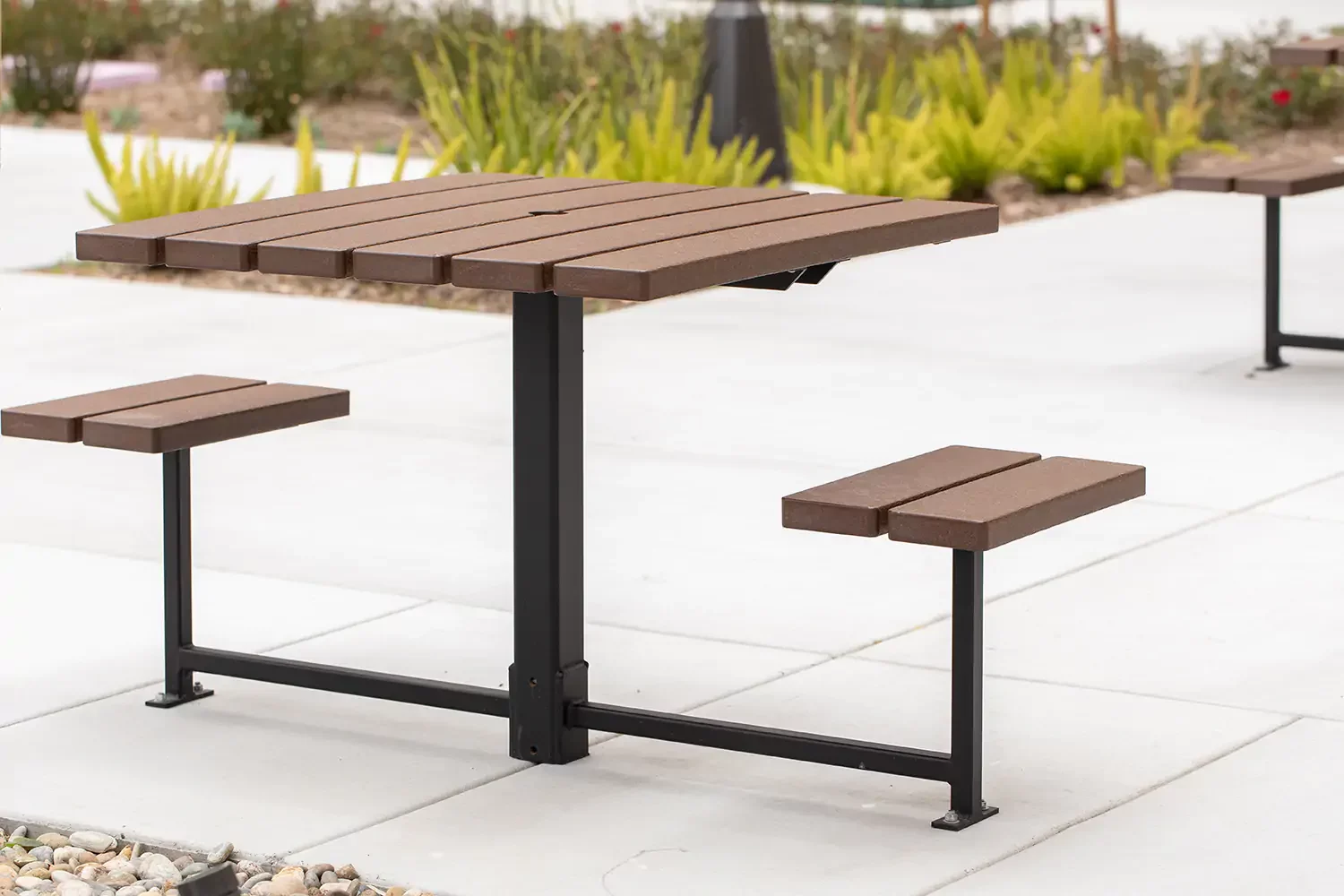 A modern outdoor picnic table with two attached benches, made of brown planks and black metal supports, set on a paved area with plants in the background.