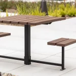 A modern outdoor picnic table with two attached benches, made of brown planks and black metal supports, set on a paved area with plants in the background.