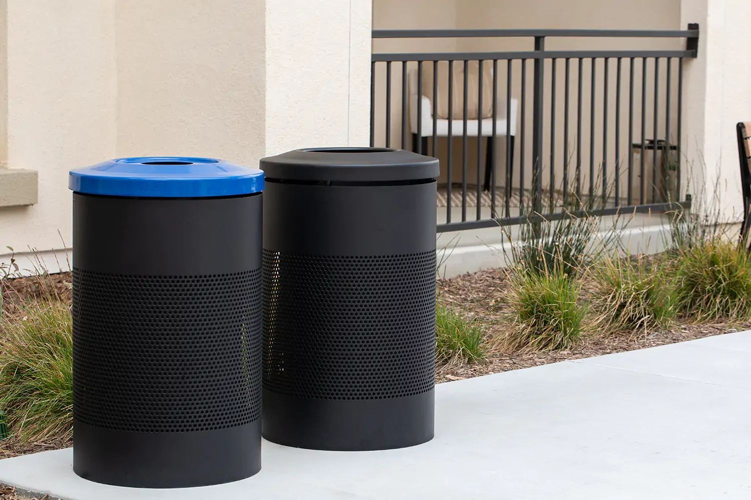 Two cylindrical outdoor trash bins, one with a blue lid and one with a black lid, stand on a sidewalk next to a building and some landscaping.