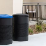 Two cylindrical outdoor trash bins, one with a blue lid and one with a black lid, stand on a sidewalk next to a building and some landscaping.