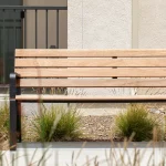 A wooden bench with black metal supports is positioned in front of a building, surrounded by ornamental grasses and landscaping.