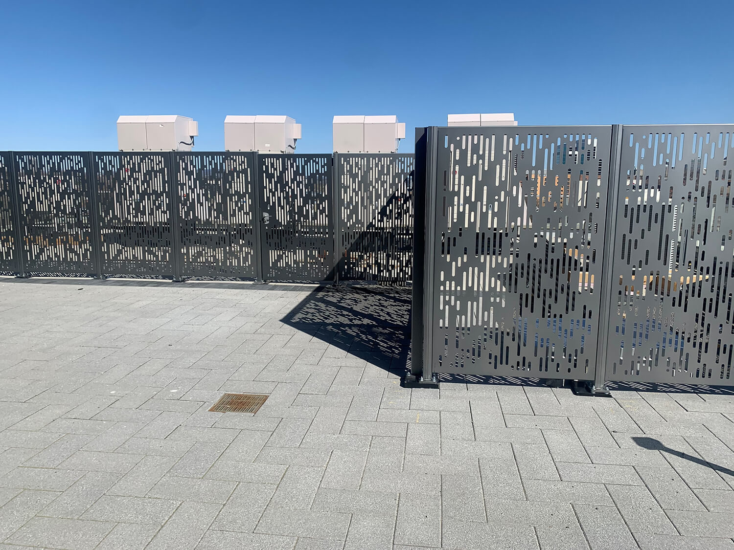 maglin_flexx-panels_6 Metal fence panels with a rectangular cutout pattern stand on a tiled surface under a clear blue sky; four box-shaped units are visible behind the fence.