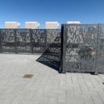 Metal fence panels with a rectangular cutout pattern stand on a tiled surface under a clear blue sky; four box-shaped units are visible behind the fence.