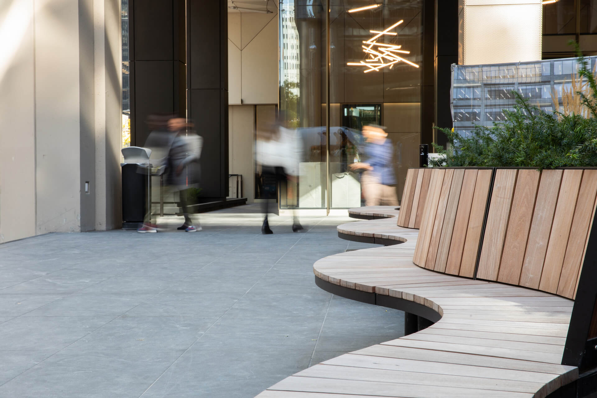 Curved wooden benches line a modern outdoor plaza with blurred figures walking past large glass doors and greenery to the right.