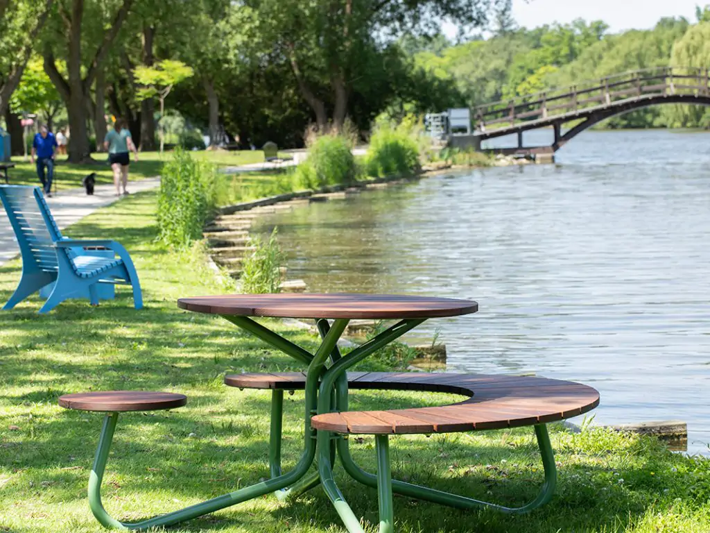 Round wooden picnic table with attached benches on grass by a lake, blue chairs nearby, people walking on a path and a wooden bridge in the background.