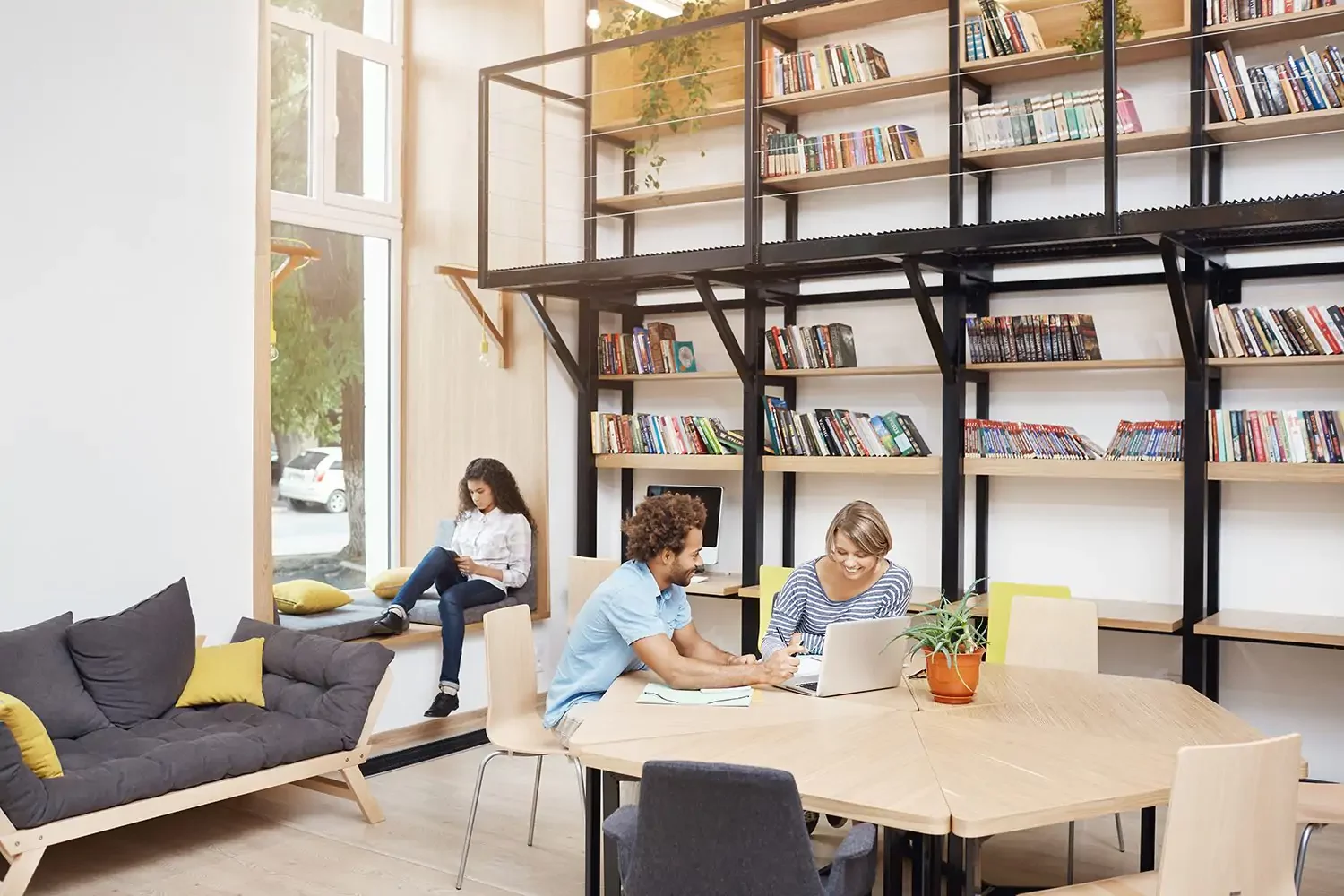 Three people are in a modern library space; two are working together at a table with a laptop, while another person is reading by the window. Shelves of books and a sofa are visible.