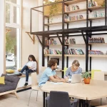 Three people are in a modern library space; two are working together at a table with a laptop, while another person is reading by the window. Shelves of books and a sofa are visible.
