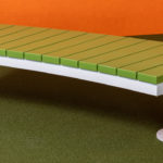 A modern bench with a green slatted seat and white metal legs, placed on green carpet with orange walls in the background.