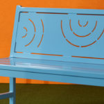 A light blue metal bench with geometric cutout patterns on the backrest, placed on green flooring with an orange background.