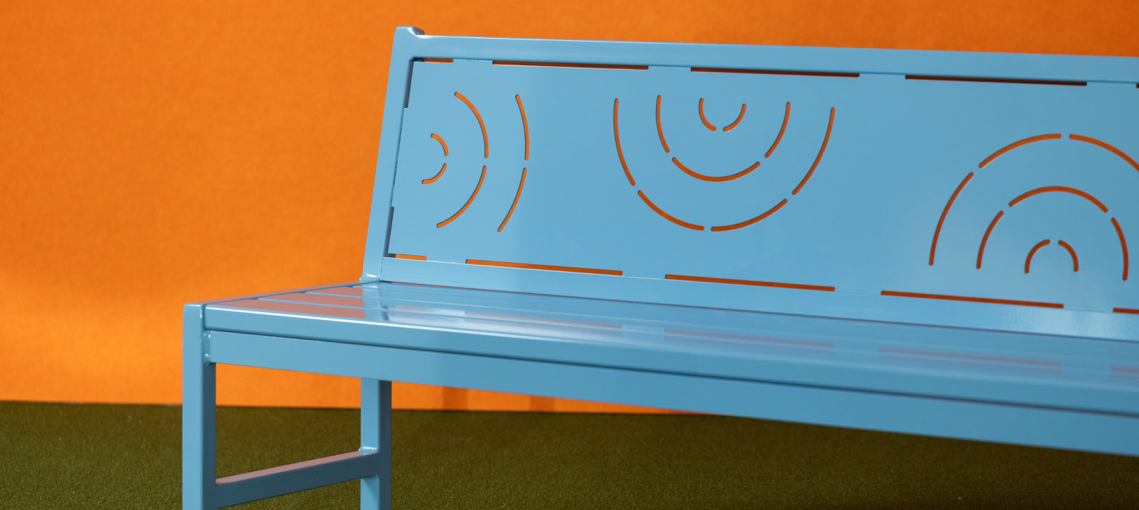 Bench2 A light blue metal bench with geometric cutout patterns on the backrest, placed on green flooring with an orange background.
