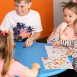 Five children sit around a small blue table, drawing and playing with toy cars. Two coloring pages are on the table, and a large plant is in the background.