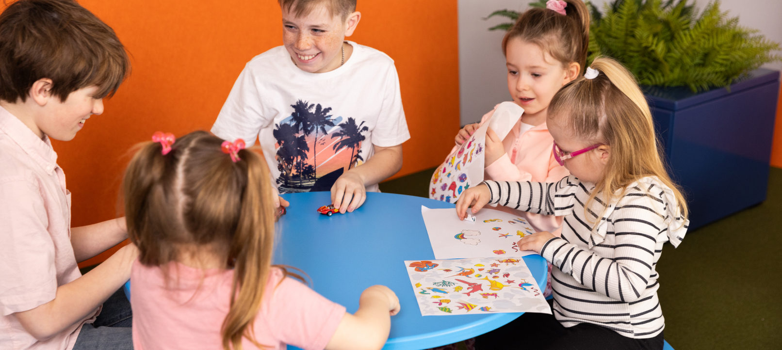 Children Five children sit around a small blue table, drawing and playing with toy cars. Two coloring pages are on the table, and a large plant is in the background.