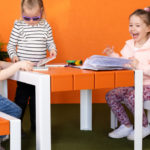 Three young children sit and stand around an orange and white table, looking at books and papers, with potted plants in the background.