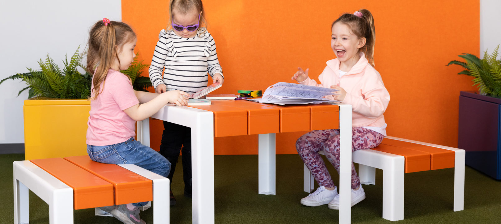 Three young children sit and stand around an orange and white table, looking at books and papers, with potted plants in the background.