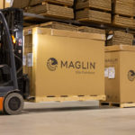 Deb drives the forklift carrying a brown Maglin box at the Maglin warehouse.