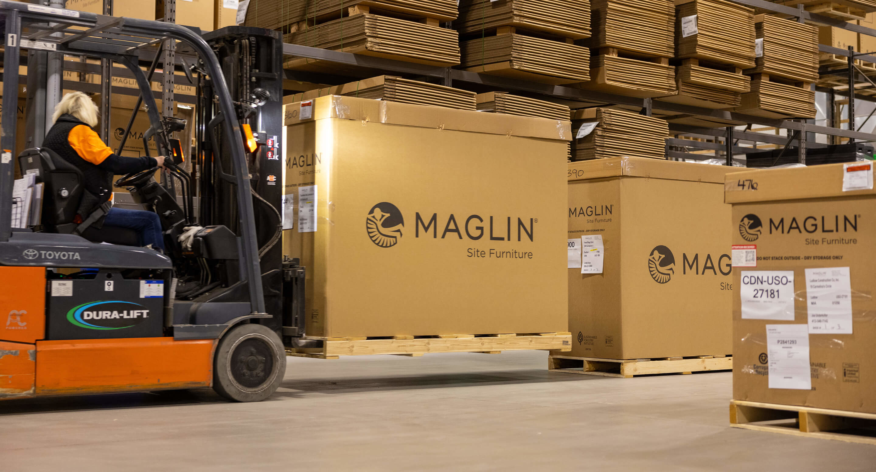 Deb drives the forklift carrying a brown Maglin box at the Maglin warehouse.