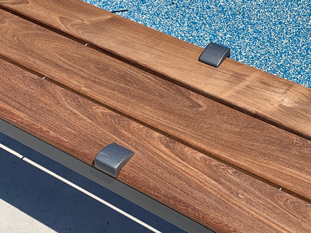 Close-up of a wooden bench with metal brackets, next to a blue speckled surface, possibly near a pool or sports area.
