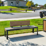 A wooden bench and trash bin are placed on a sidewalk near a grassy area, with motorcycles parked and buildings visible in the background.