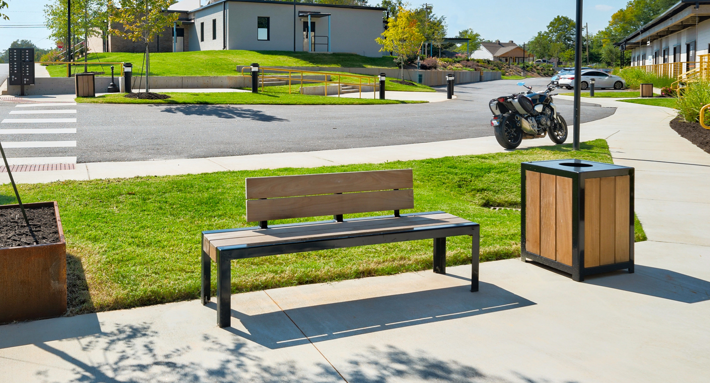 maglin-collections-1050-gallery A wooden bench and trash bin are placed on a sidewalk near a grassy area, with motorcycles parked and buildings visible in the background.