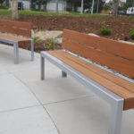 Two modern wooden benches with metal frames are placed on a concrete surface in an outdoor park area with mulch, stones, and small plants.