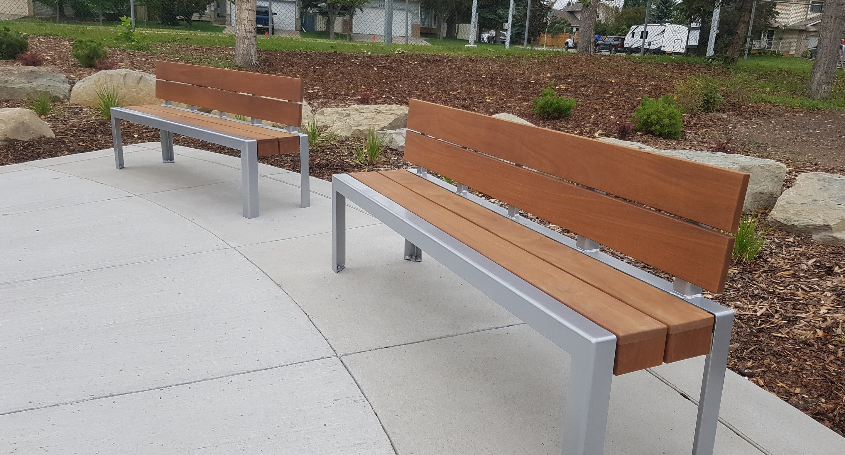 maglin-collections-1050-gallery2 Two modern wooden benches with metal frames are placed on a concrete surface in an outdoor park area with mulch, stones, and small plants.