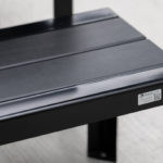 Close-up of a black metal bench with a small label on the side, positioned on a light-colored surface.