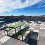 Rooftop patio with checkered tile floor, colorful picnic tables, lounge chairs, and a city skyline in the background under a blue sky with wispy clouds.